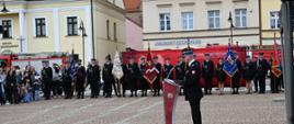 Na pierwszym planie, tyłem do obiektywu, stoi strażak w ciemnym mundurze galowym, który przemawia przy nowoczesnej, czerwonej mównicy z logo Państwowej Straży Pożarnej. W tle, na wprost przemawiającego, ustawione są rzędy strażaków oraz poczty sztandarowe z ozdobnymi sztandarami w różnych kolorach. Za nimi widoczna jest duża czerwona ścianka z napisem „Państwowa Straż Pożarna”, a w oddali zabytkowe kamienice oraz zgromadzeni widzowie, w tym rodziny z dziećmi.