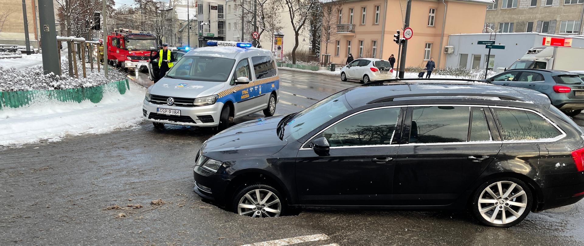 Skrzyżowanie Alei Niepodległości z ul. Kochanowskiego Samochód osobowy kombi wpadł lewym kołem do powstałej dziury i zawisł na podłodze. Za samochodem ardiowóz Strazy Miejskiej. Policjant a dalej samochody straży pożarnej