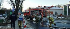 Na ulicy stoi samochód pożarniczy. Strażak PSP i strażak OSP zwijają węże tłoczne. Po lewej stronie kierujący działaniem ratowniczym wraz z funkcjonariuszem Policji idą chodnikiem w stronę miejsca zdarzenia. W tle na przeciwległej stronie ulicy przedstawiciele zarządzania kryzysowego.