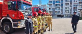 Komendant główny Państwowej Straży Pożarnej wita się ze strażakami stojącymi w rzędzie za nimi stoją samochody ratowniczo-gaśnicze.