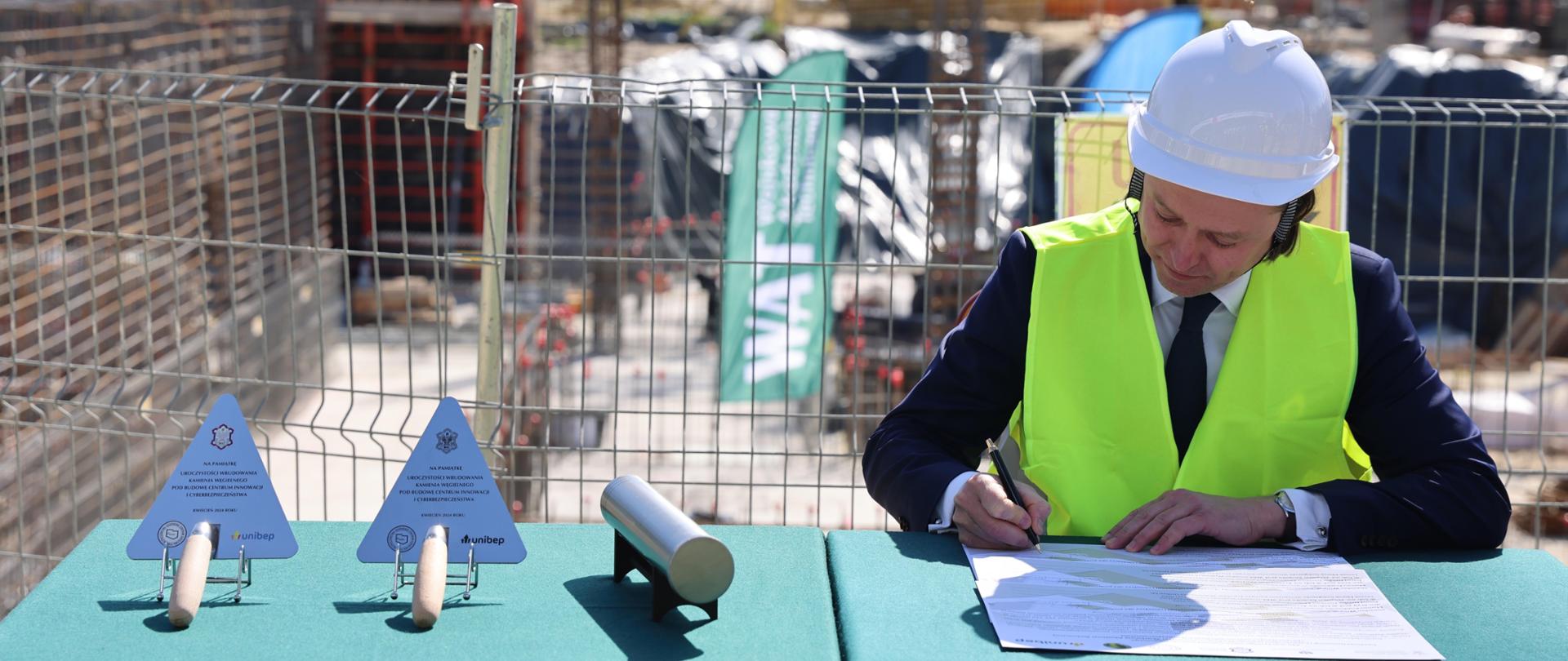 Na zdjęciu mężczyzna podpisuje oficjalny akt wmurowania. Ma na sobie żółtą kamizelkę oraz biały kask. 