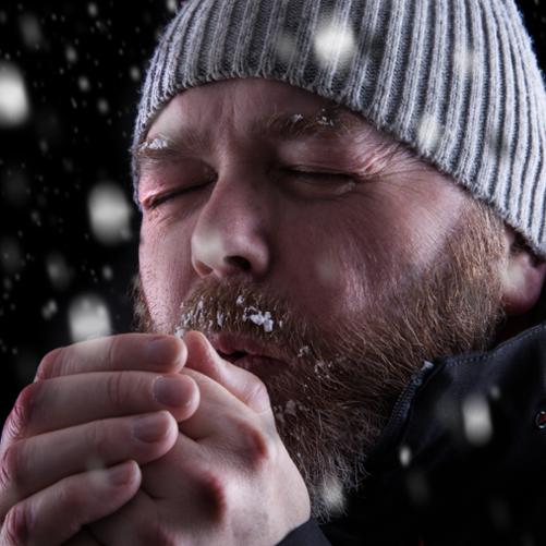 Marznący brodaty mężczyzna w czapce chucha na zmarznięte zaczerwienione ręce. Wokół jest ciemno i pada śnieg.