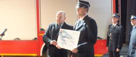 Wręczenie promesy w Jutrosinie. Plac przed remizą OSP w Jutrosinie. Minister Jan Dziedziczak wręczył promesę prezesowi OSP w Jutrosinie. Obaj na tle wozu strażackiego pozują do zdjęcia. Za nimi widocznych kilku innych uczestników spotkania.