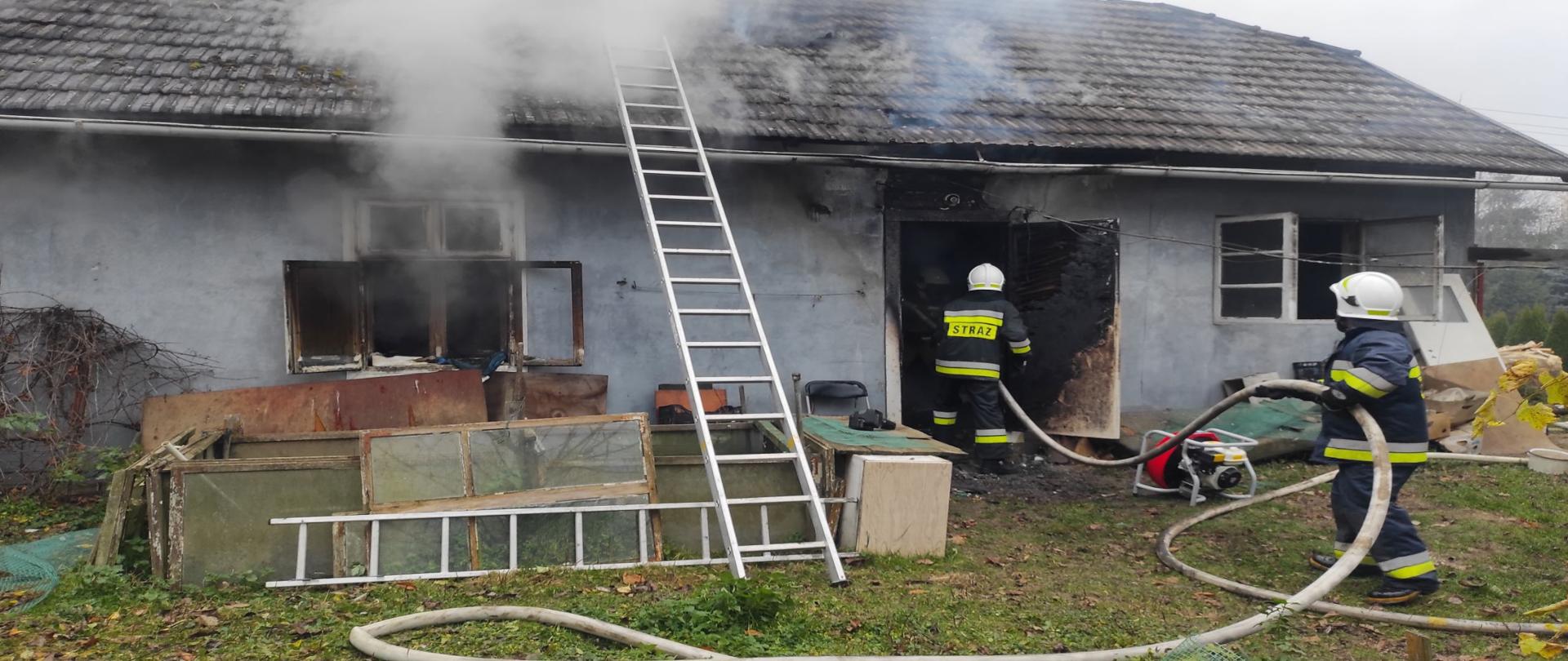 Kolorowa fotografia przedstawia akcję gaśniczą straży pożarnej gaszenia drewnianego budynku mieszkalnego. Na pierwszym planie dwóch strażaków w ubraniach bojowych, w hełmach na głowach uczestniczą w akcji gaśniczej. Przed lekko nadpalonym budynkiem widoczne białe węże pożarnicze wykorzystywane go podawania wody na pożaru wewnętrznego.