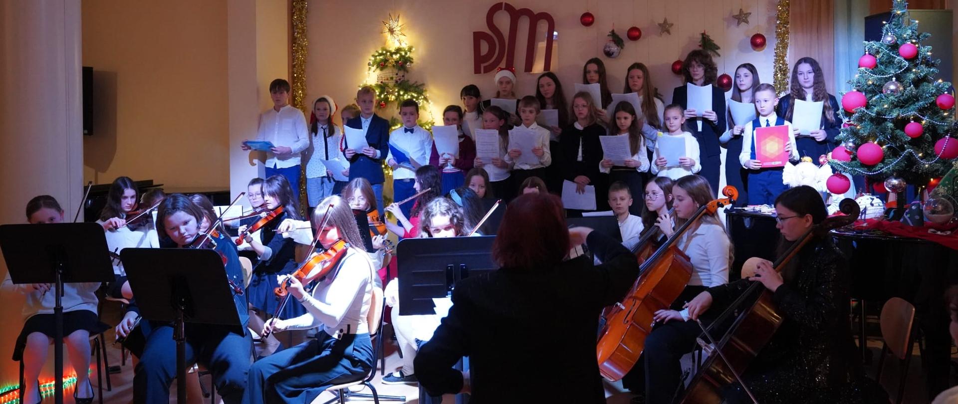 Na dole zdjęcia na samym środku na pierwszym planie pani dyrygent dalej widoczna orkiestra grająca na różnych instrumentach między innymi widoczne skrzypce i wiolonczele. Za orkiestrą na podwyższeniu stoi chór. Za chórem na ścianie widoczne ozdoby świąteczne oraz logo szkoły PSM. Po prawej stronie na fortepianie widoczne ozdoby świąteczne oraz choinka