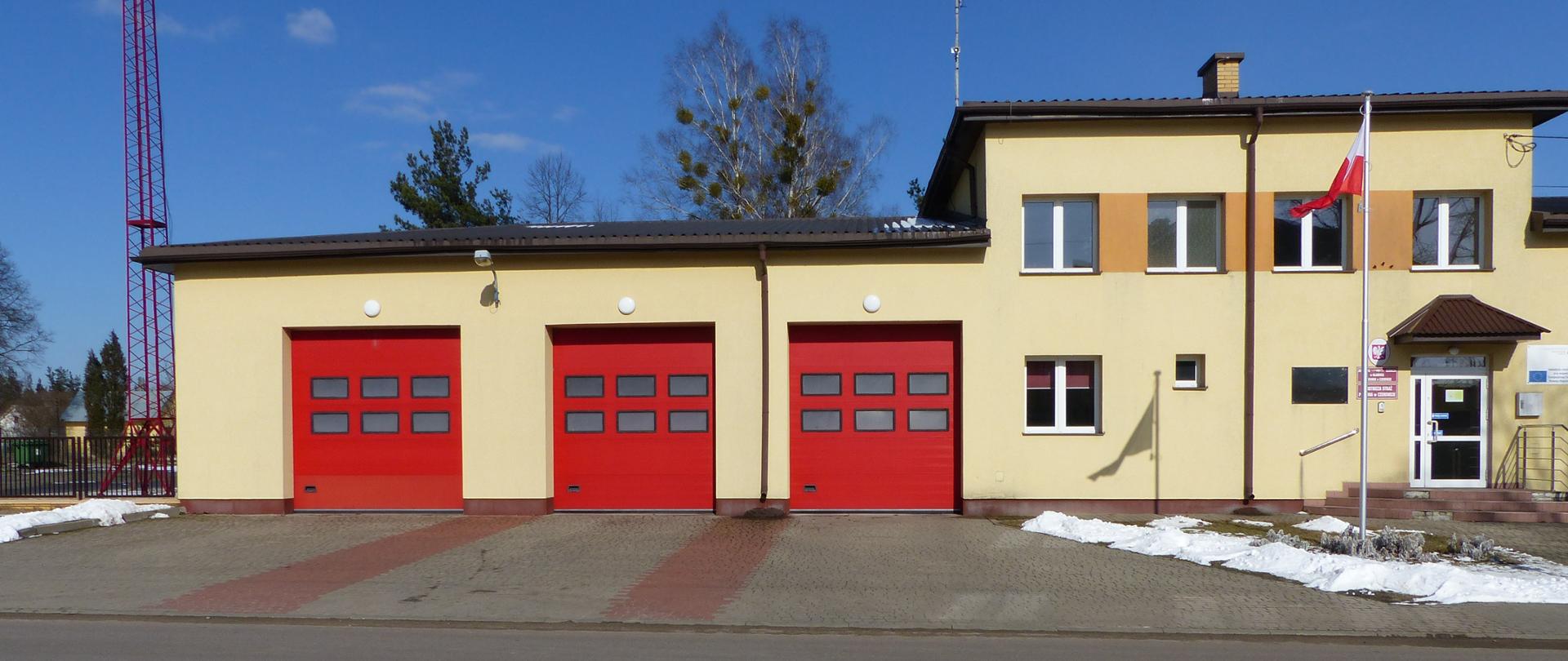 Budynek Posterunku Jednostki Ratowniczo Gaśniczej w Czeremsze. Budynek z trzema czerwonymi bramami garażowymi. Elewacja budynku w kolorze żółtym. Przed budynkiem za maszcie powiewa Flaga Państwowa.