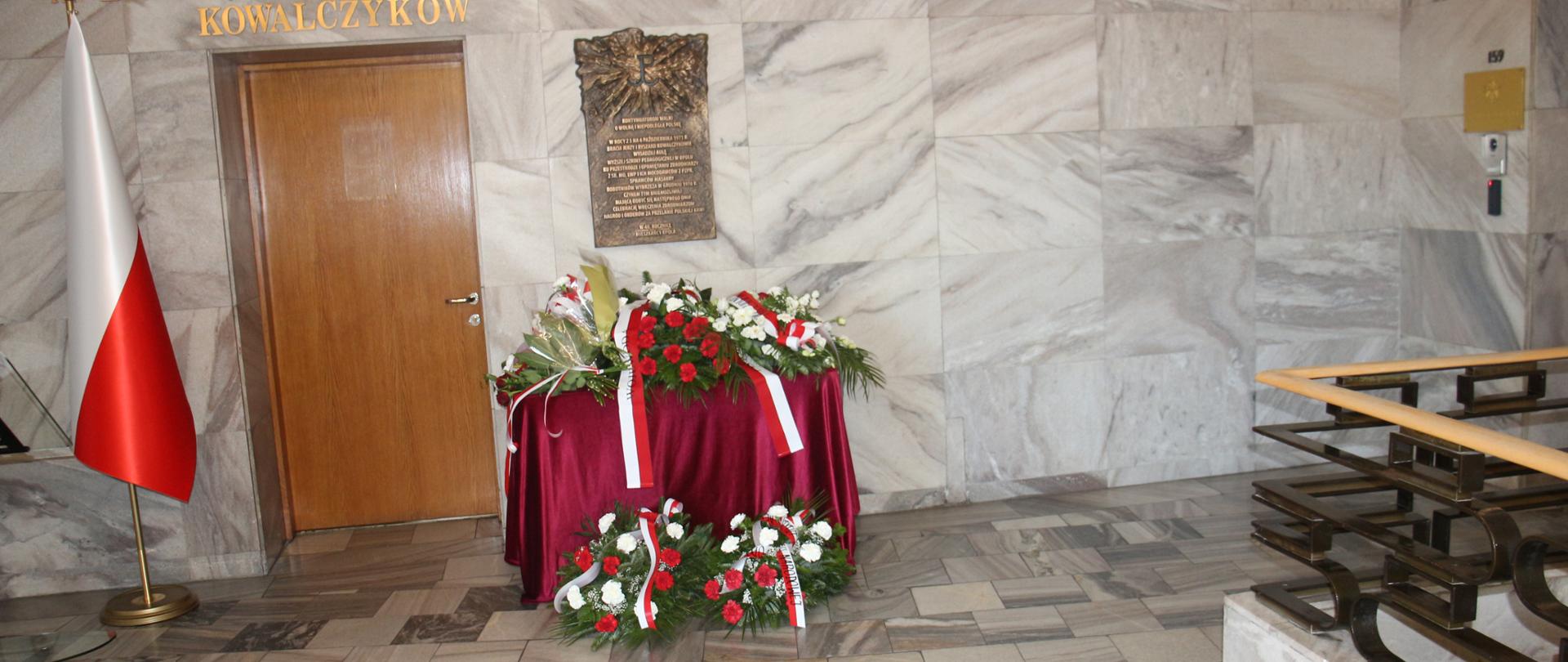Po lewej stronie polska flaga, dalej brązowe drzwi, nad nimi napis ""Sala braci Jerzego i Ryszarda Kowalczyków". Obok na ścianie bazowa tablica pamiątkowa pod nią złożone kwiaty.
