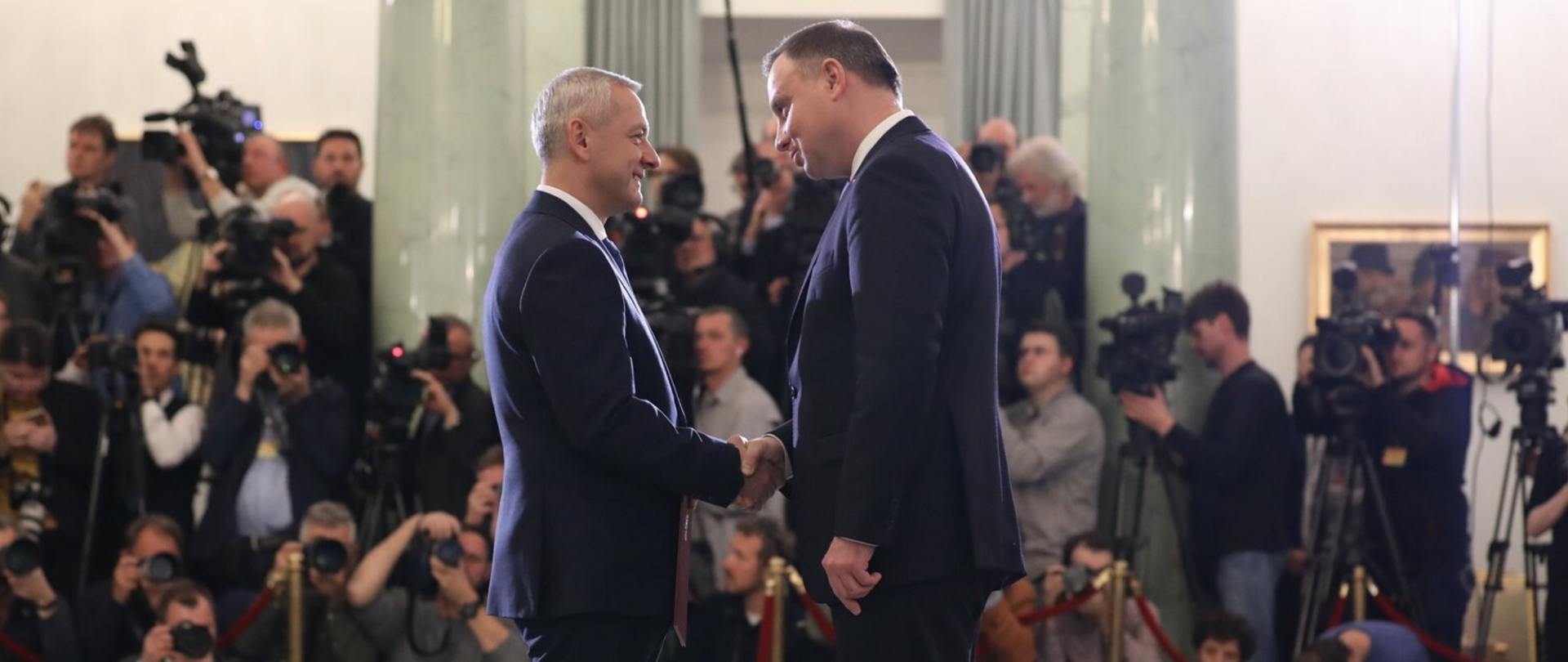 Minister Marek Zagórski i prezydent Andrzej Duda podają sobie ręce. W tle tłum przedstawicieli mediów.