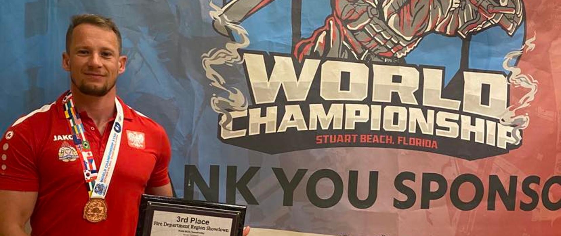 na kolorowym tle z napisami "world championship" z lewej strony stoi na czerwono ubrany mężczyzna, na szyi ma medal a w rekach na wysokości prawego biodra trzyma tablice z napisami.