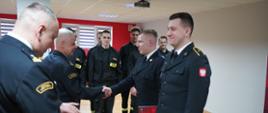 Świetlica Komendy Powiatowej PSP w Rawiczu. Zastępca komendanta powiatowego PSP ściskając dłoń strażaka, składa mu gratulacje z okazji awansu na wyższe stanowisko służbowe. W tle pozostali uczestnicy uroczystej zbiórki. 