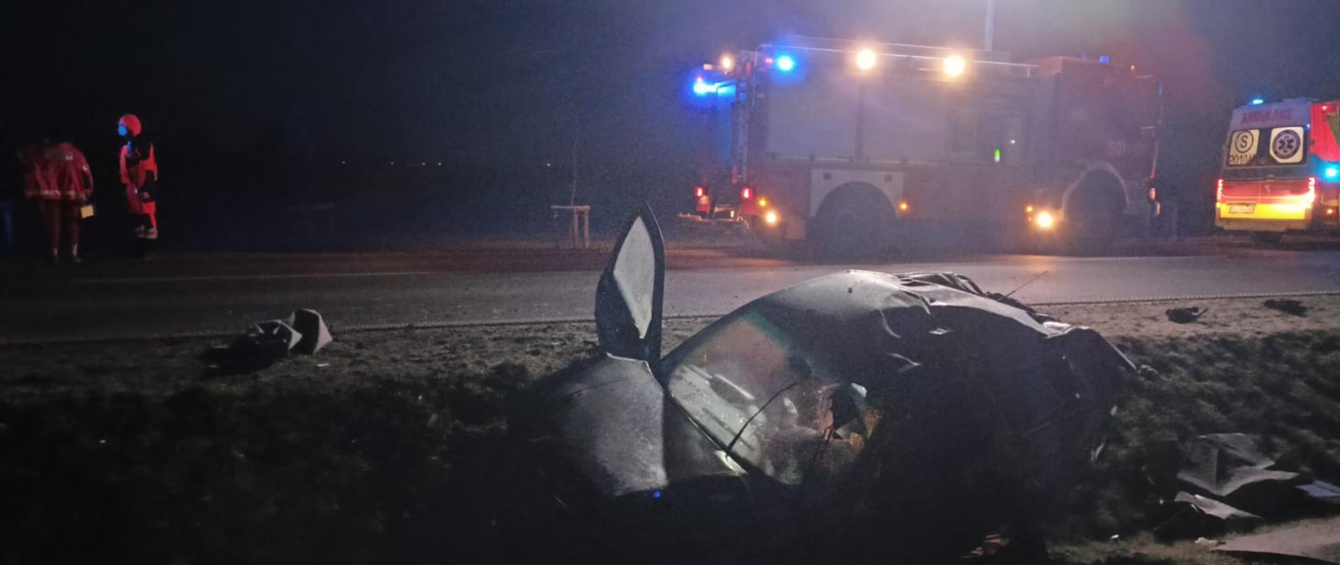 Widać samochód osobowy w rowie, jest ciemno, w oddali samochód strażacki