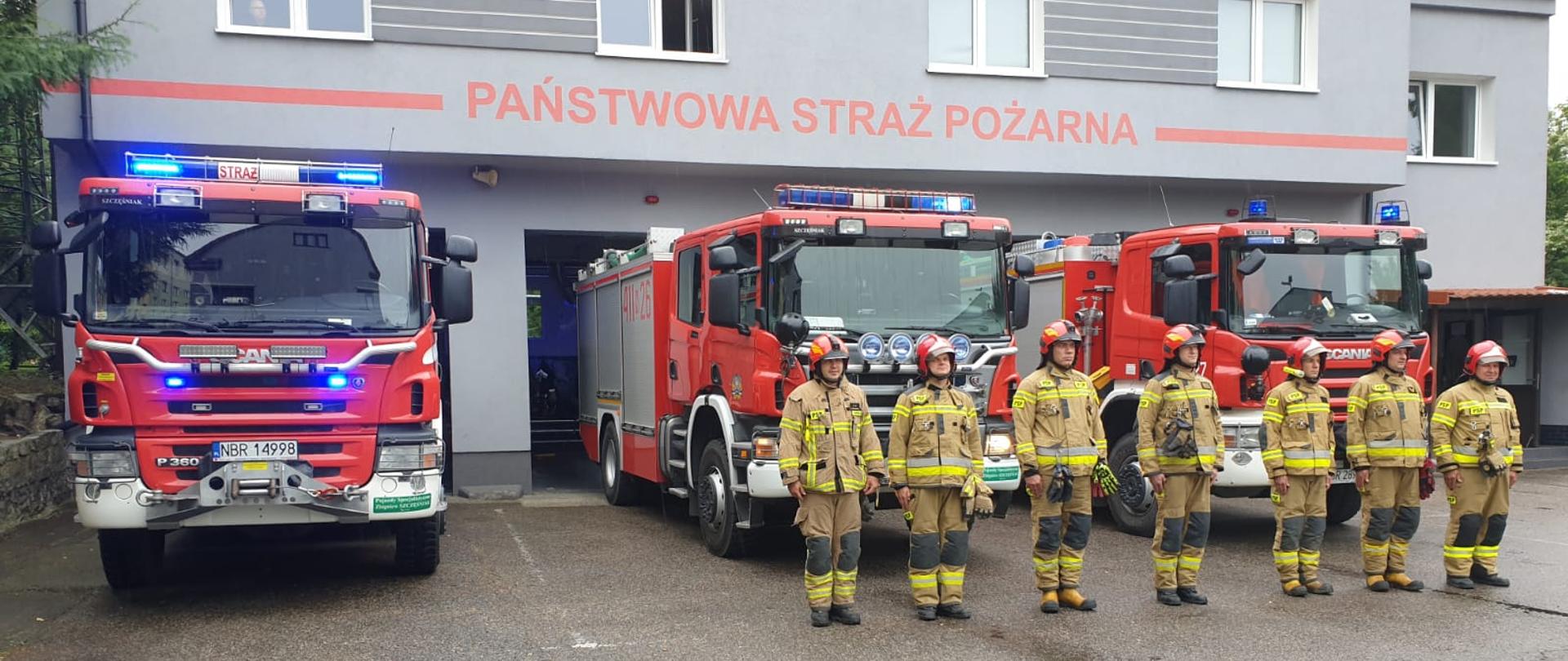Strażacy w mundurach stoją przy czerwonych samochodach, z tyłu strażnica.