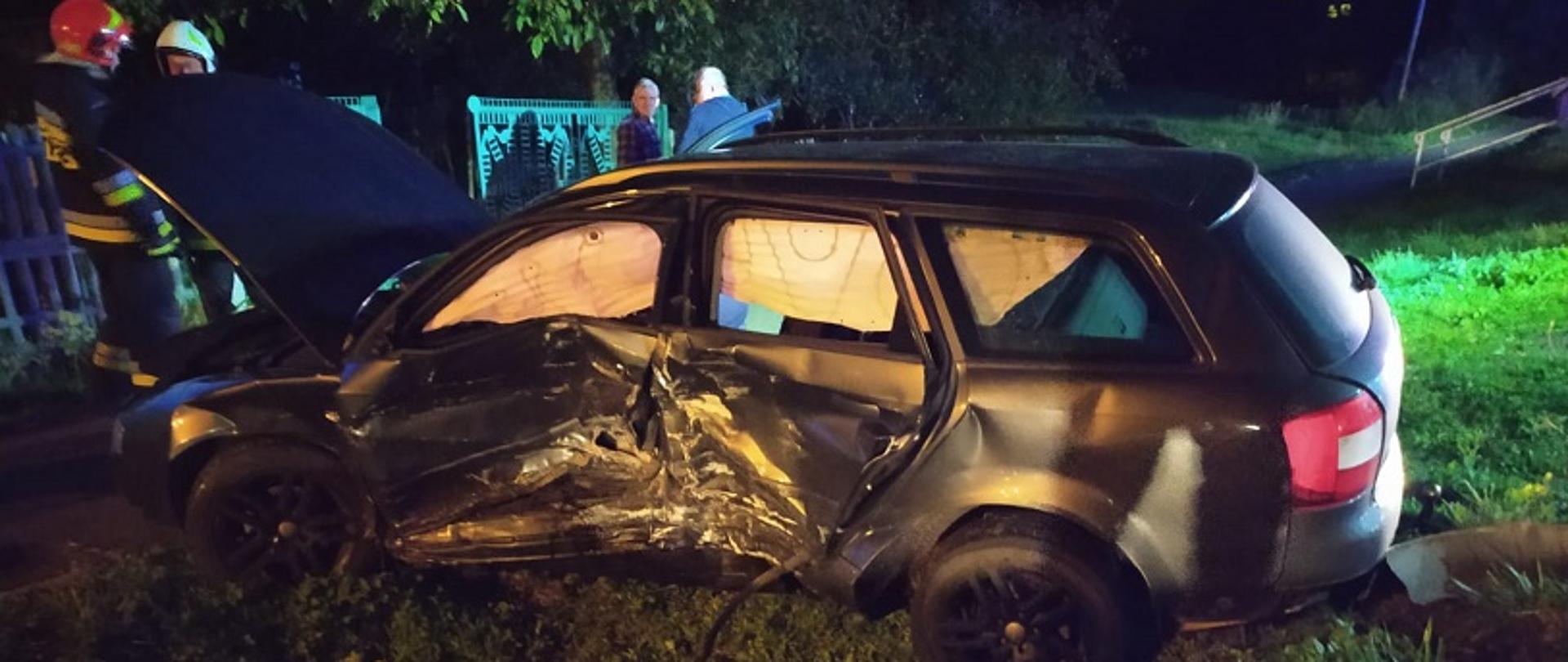 Zdjęcie przedstawia samochód osobowy, który w wyniku zderzenia znalazł się na poboczu drogi. Samochód ma rozbity lewy bok.