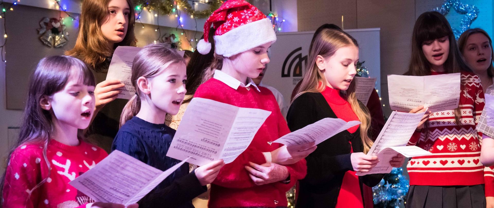 zdjęcie przedstawia grupę uczennic klas młodszych śpiewających z nut na scenie sali koncertowej, w tle baner szkoły i dekoracje świąteczne