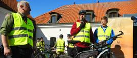 Minister Czarnek w żółtej kamizelce stoi przy rowerze i mówi do trzymanego w ręku mikrofonu, obok niego kilka osób w takich samych kamizelkach, w tle stary biały budynek z dachem krytym czerwoną dachówką.