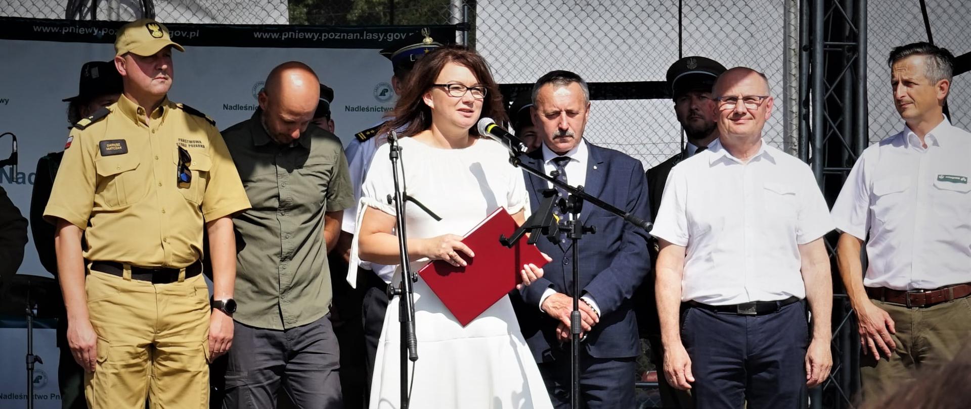grupa osób stoi na podwyższeniu, kobieta w białej sukni mówi do mikrofonu trzymającczerwoną teczkę, za nią stoją mężczyźni, jeden z nich jest w mundurze