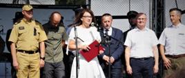 grupa osób stoi na podwyższeniu, kobieta w białej sukni mówi do mikrofonu trzymającczerwoną teczkę, za nią stoją mężczyźni, jeden z nich jest w mundurze