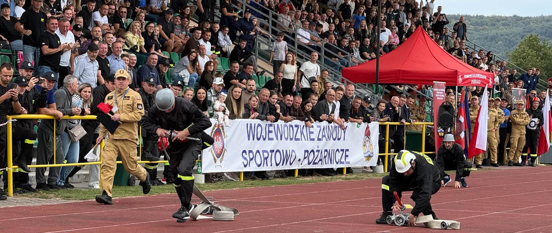 Strażacy wykonują konkurencję bojową na czerwonej bieżni lekkoatletycznej podczas Wojewódzkich Zawodów Sportowo-Pożarniczych przed pełnymi widowni trybunami stadionu.