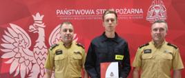Na zdjęciu widnieje nowy funkcjonariusz z komendantem miejskim PSP w Koninie oraz zastępcą komendanta. W tle czerwony baner KM PSP w Koninie.