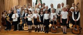 Grupa dziecięca pozuje do wspólnego zdjęcia na scenie, trzymając różnorodne instrumenty muzyczne, m.in. gitary, skrzypce, flet i instrumenty dęte. Wszyscy są odświętnie ubrani, a w tle widać bożonarodzeniowe dekoracje.