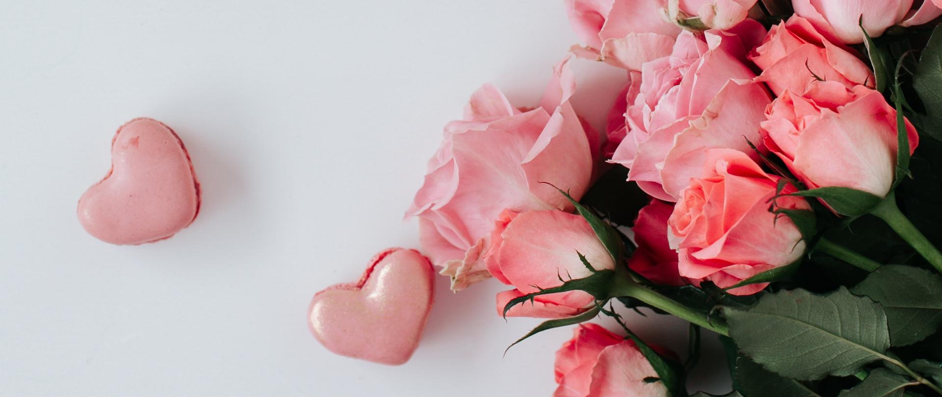 z prawej strony bukiet różowych róż, z lewej strony oraz powyżej bukietu różowe serca, w centralnej części filiżanka kawy z pianą z mleka widoczna od góry