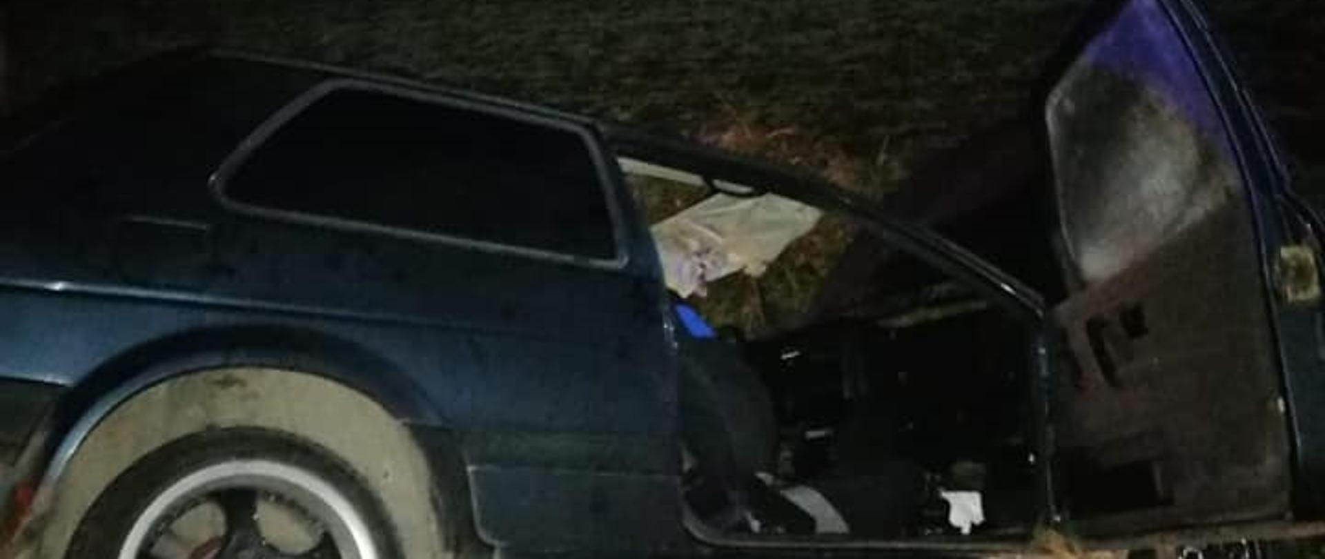 Widać ciemny samochód z otwartymi drzwiami od pasażera z prawej strony, który jest w rowie