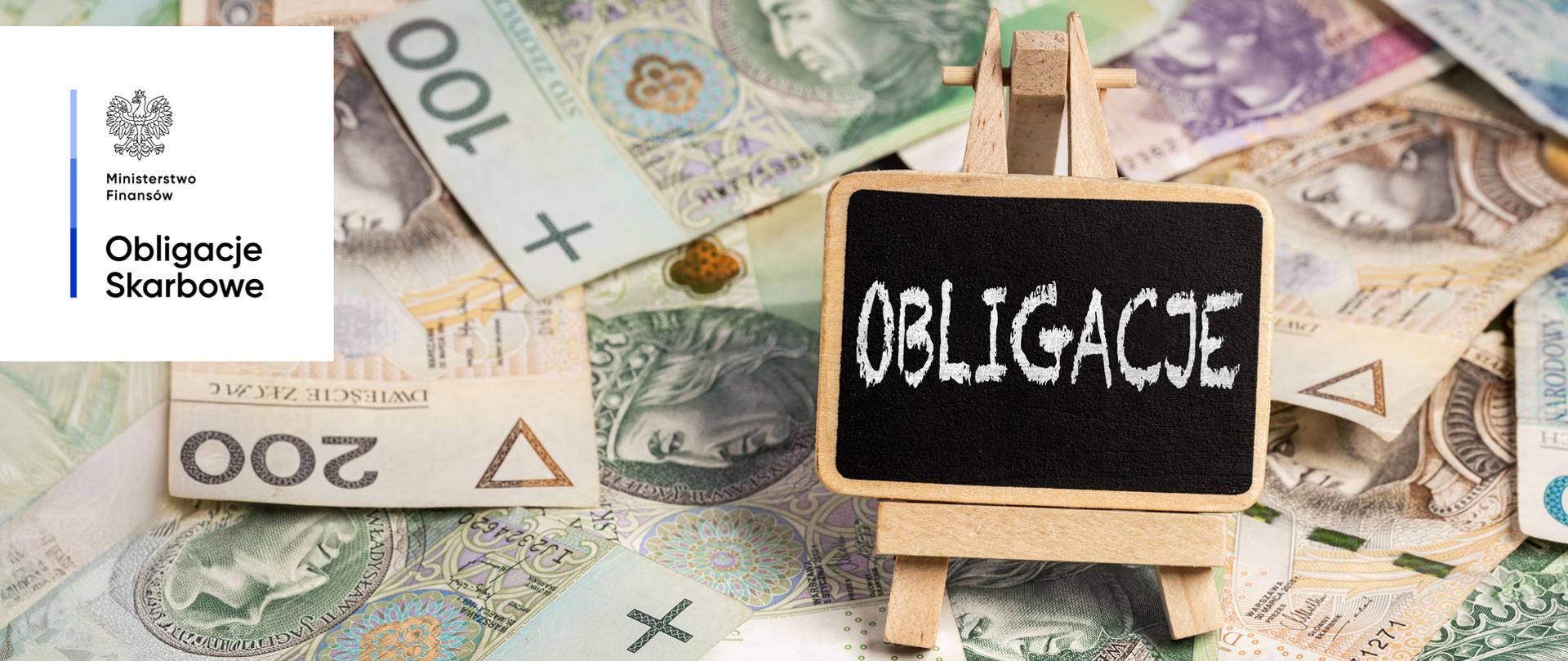 Tabliczka z napisem "Obligacje" ustawiona na sztaludze, otoczona rozłożonymi polskimi banknotami o różnych nominałach. Ministerstwo Finansów, Obligacje Skarbowe.