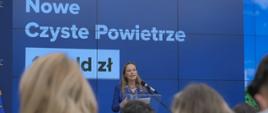 Minister funduszy i polityki regionalnej Katarzyna Pełczyńska-Nałęcz przedstawia założenia rewizji KPO, minister stoi przy pulpicie z mikrofonem, za jej plecami grafika z niebieskim tłem i informacją o 14 mld zł na nowe Czyste Powietrze