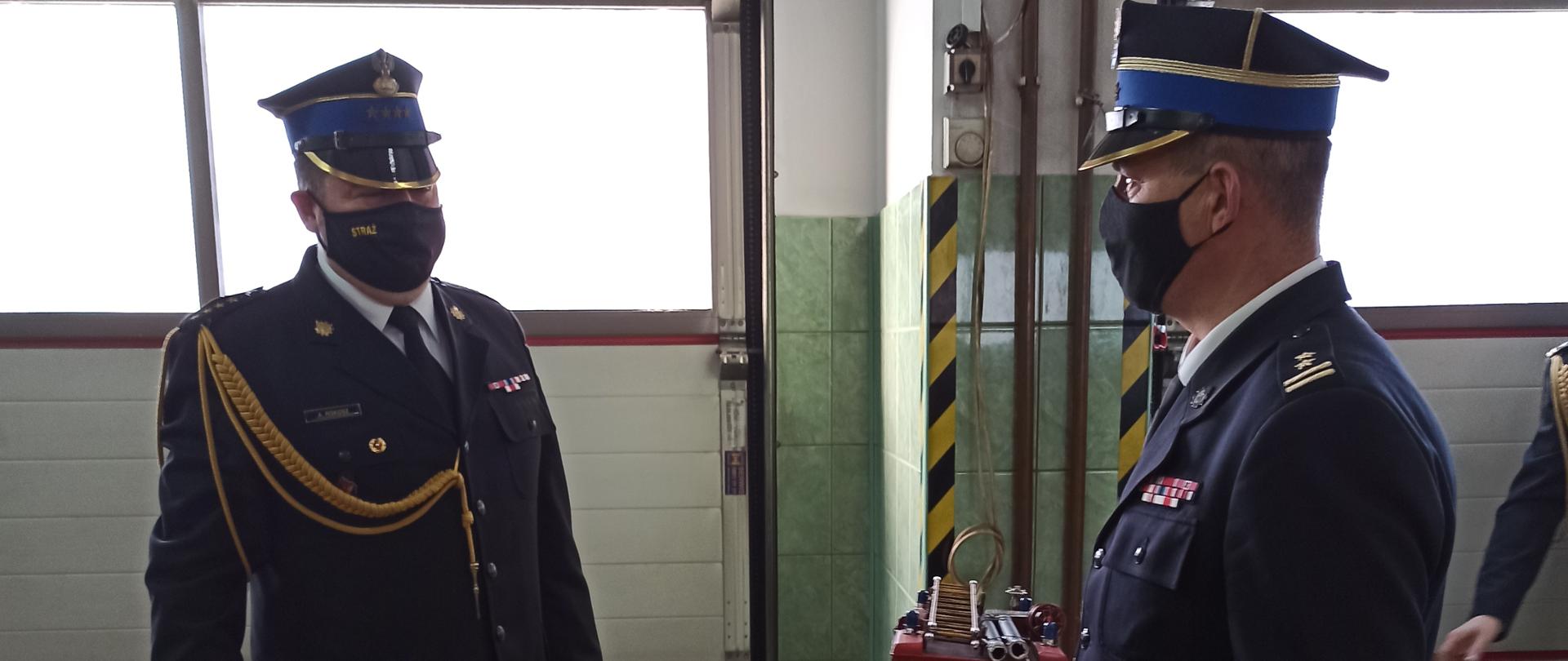 zdjęcie zrobione zostało w garażu JRG Rabka-Zdrój na zdjęciu znajdują się komendant powiatowy w granatowym mundurze w ręku trzyma replikę samochodu pożarniczego i wręcza go strażakowi który przechodzi na emeryturę ubranemu w mundur wyjściowy granatowy ze sznurem i baretkami