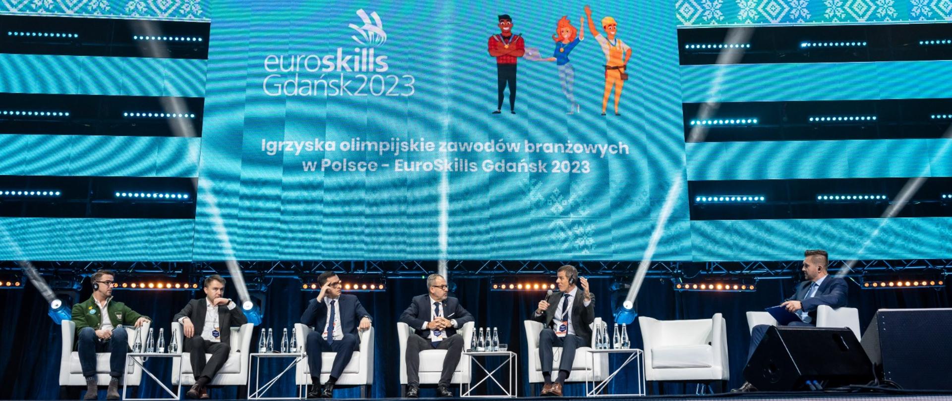 Na scenie 5 prelegentów i jeden prowadzący. Za nimi wyświetlana prezentacja na dużych telebimach z napisem: Igrzyska olimpijskie zawodów branżowych w Polsce - EuroskillsGdańsk2023. 
