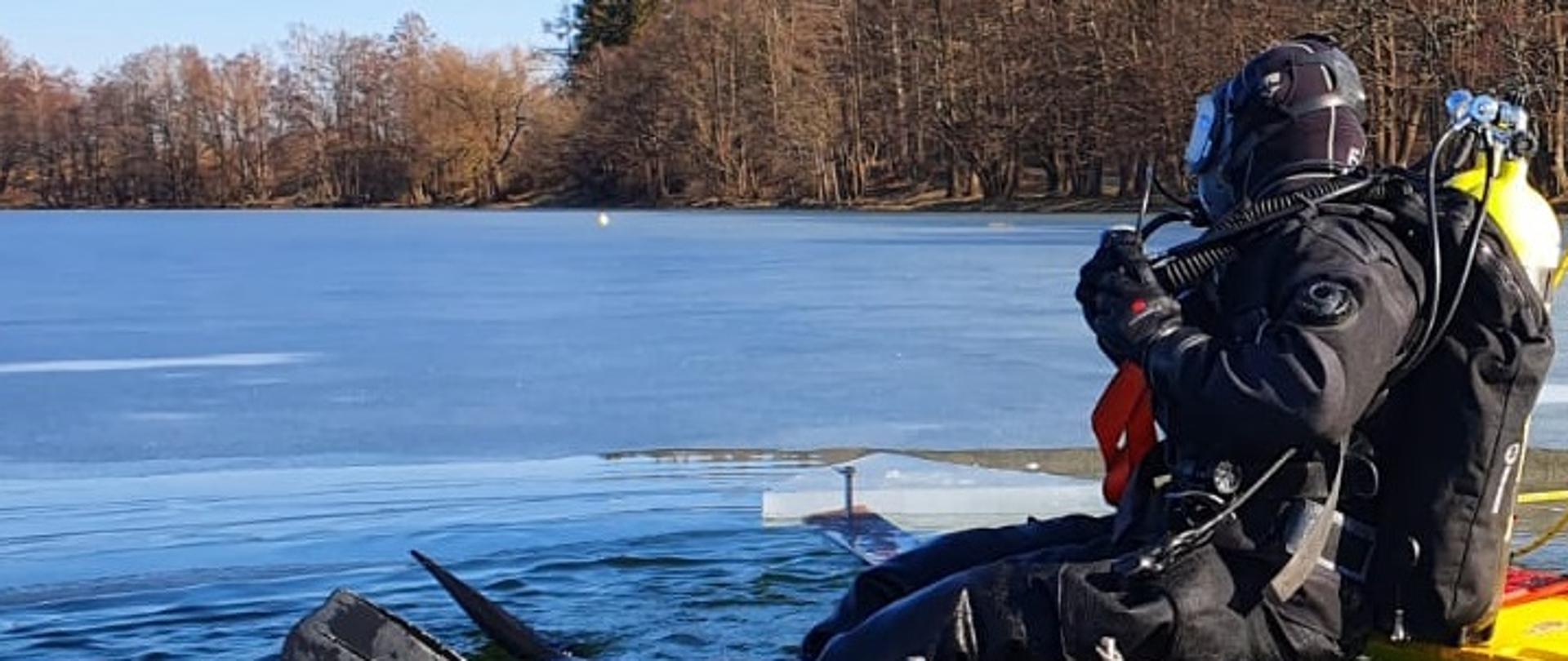 Nurek ubrany w sprzęt nurkowy siedzi na lodzie przy przerębli i szykuje się do wejścia pod wodę. Za nim w oddali widać drzewa. 
