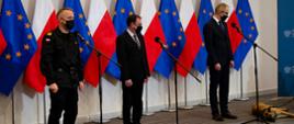 Na zdjęciu na tle biało czerwonych i unijnych flag Minister Spraw Wewnętrznych i Administracji w środku wiceminister z prawej strony i Komendant Główny Państwowej Straży Pożarnej z lewej strony. Wszyscy stoją przed mikrofonami. Obok wiceministra leży pies.