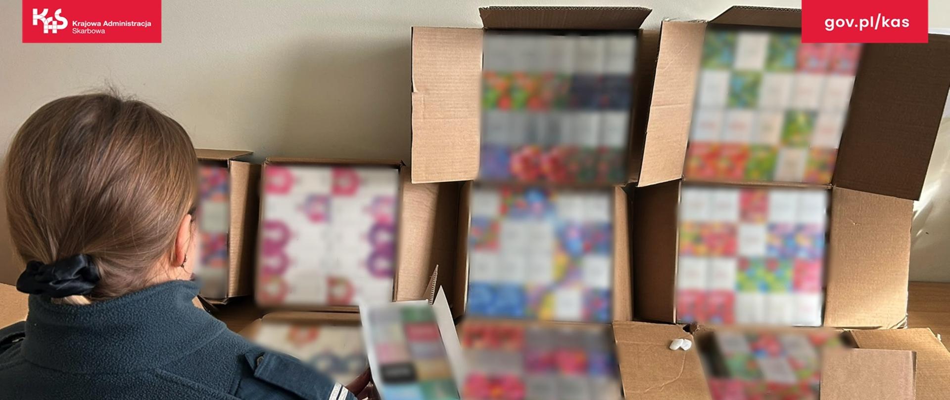 Funkcjonariuszka SCS stoi tyłem, przed nią na stole leżą kartony otwarte. W kartonach znajduje się duża ilość opakowań z liquidami i płynami do e-papierosów.