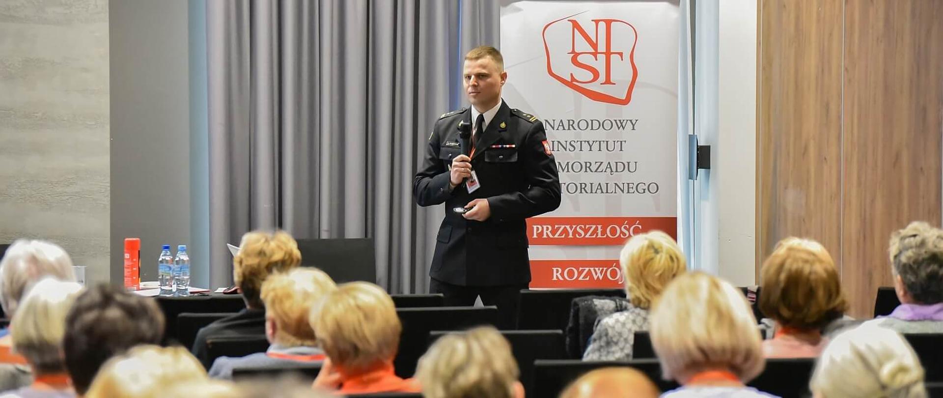 brygadier Mikołaj Pawełczyk w trakcie wykładu na sali konferencyjnej, ubrany jest w mundur wyjściowy za nim znajduje się rollup narodowego instytutu samorządu terytorialnego, na zdjęciu widać również głowy osób słuchających wykładu