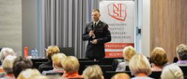 brygadier Mikołaj Pawełczyk w trakcie wykładu na sali konferencyjnej, ubrany jest w mundur wyjściowy za nim znajduje się rollup narodowego instytutu samorządu terytorialnego, na zdjęciu widać również głowy osób słuchających wykładu