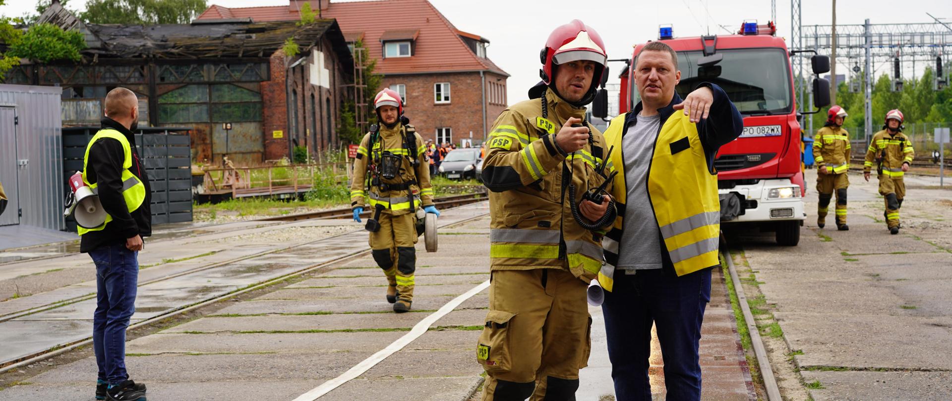 bocznica kolejowa. strażak rozmawia z osobą z PKP, z tyłu pojazd pożarniczy