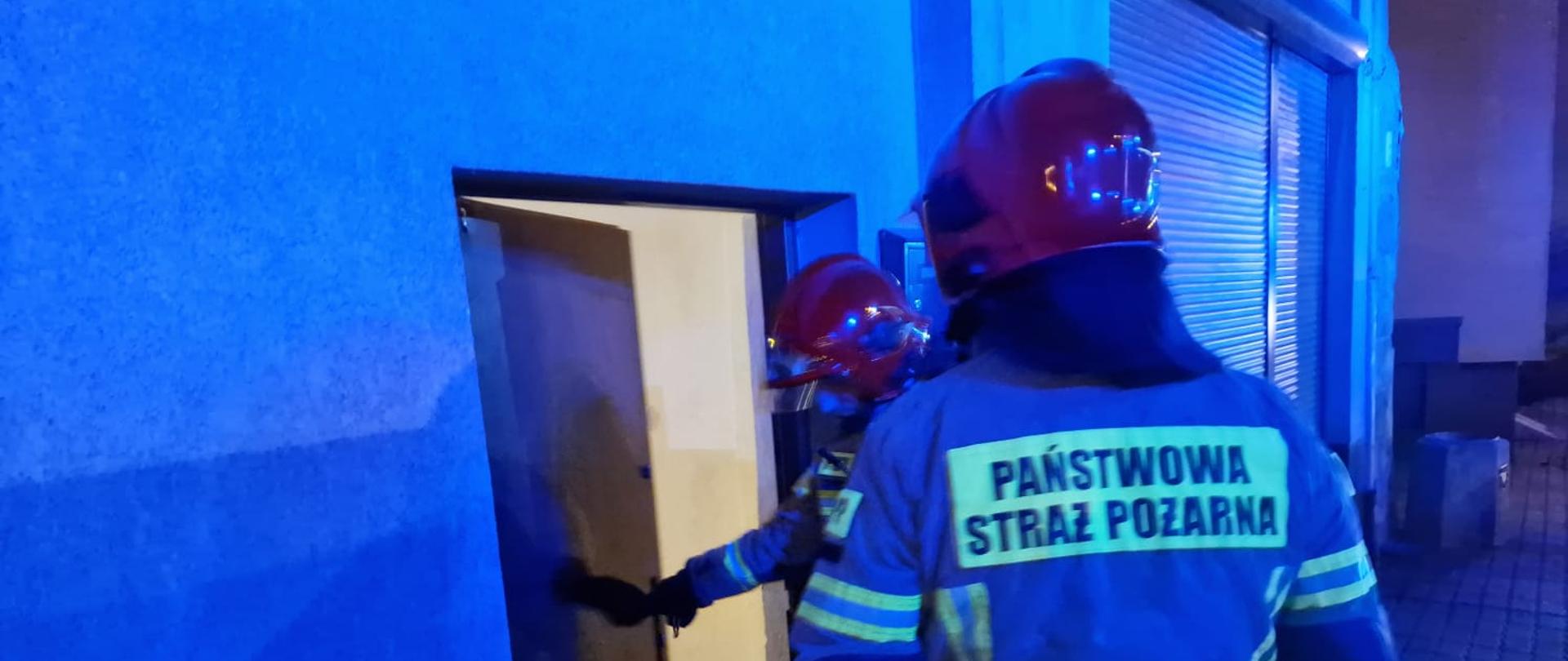Obraz przedstawia strażaków w umundurowaniu specjalnym wchodzących do budynku. Pora nocna.