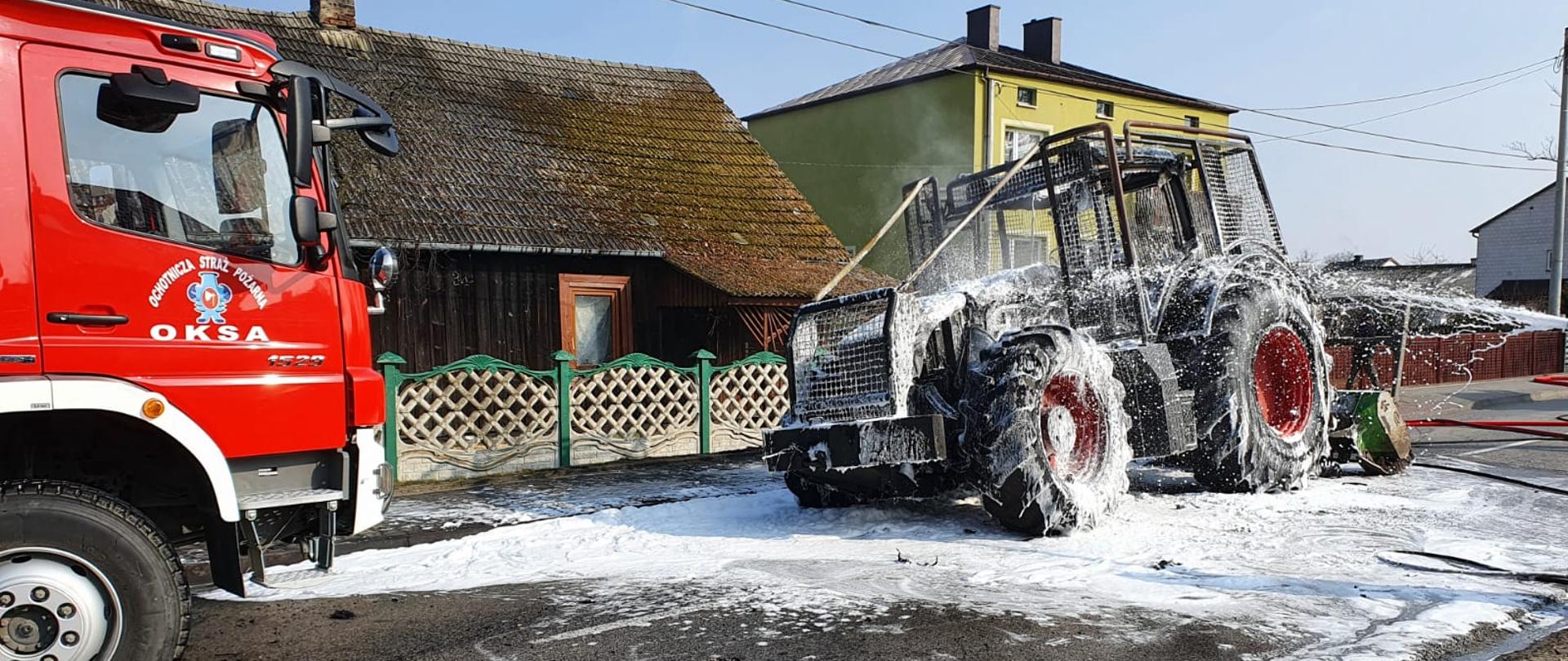Zdjęcie po lewej stronie przedstawia przód samochodu ratowniczo-gaśniczego. Na jego drzwiach widniej napis „OSP Oksa”. W środkowej części zdjęcia widać spalony ciągnik rolniczy pokryty warstwą piany gaśniczej. Zarówno pojazd strażacki jak i ciągnik znajdują się na pasie jezdni drogi wojewódzkiej. W tle widać parterowy, drewniany budynek mieszkalny. 