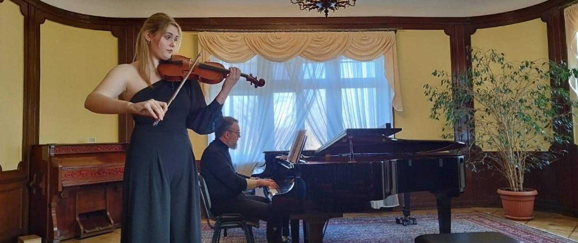 Zdjęcie kolorowe. Kobieta grająca na skrzypcach stoi w salonie o wystroju klasycznym. Ma na sobie czarny kombinezon na jedno ramię. W tle mężczyzna siedzi przy czarnym fortepianie, trzymając dłonie na klawiaturze. Pomieszczenie ma żółte ściany z ciemną, drewnianą lamperią, ozdobny żyrandol pod sufitem oraz duże okno z jasnymi firankami i zasłonami. Po prawej stronie stoi roślina doniczkowa, a podłogę pokrywa wzorzysty dywan.