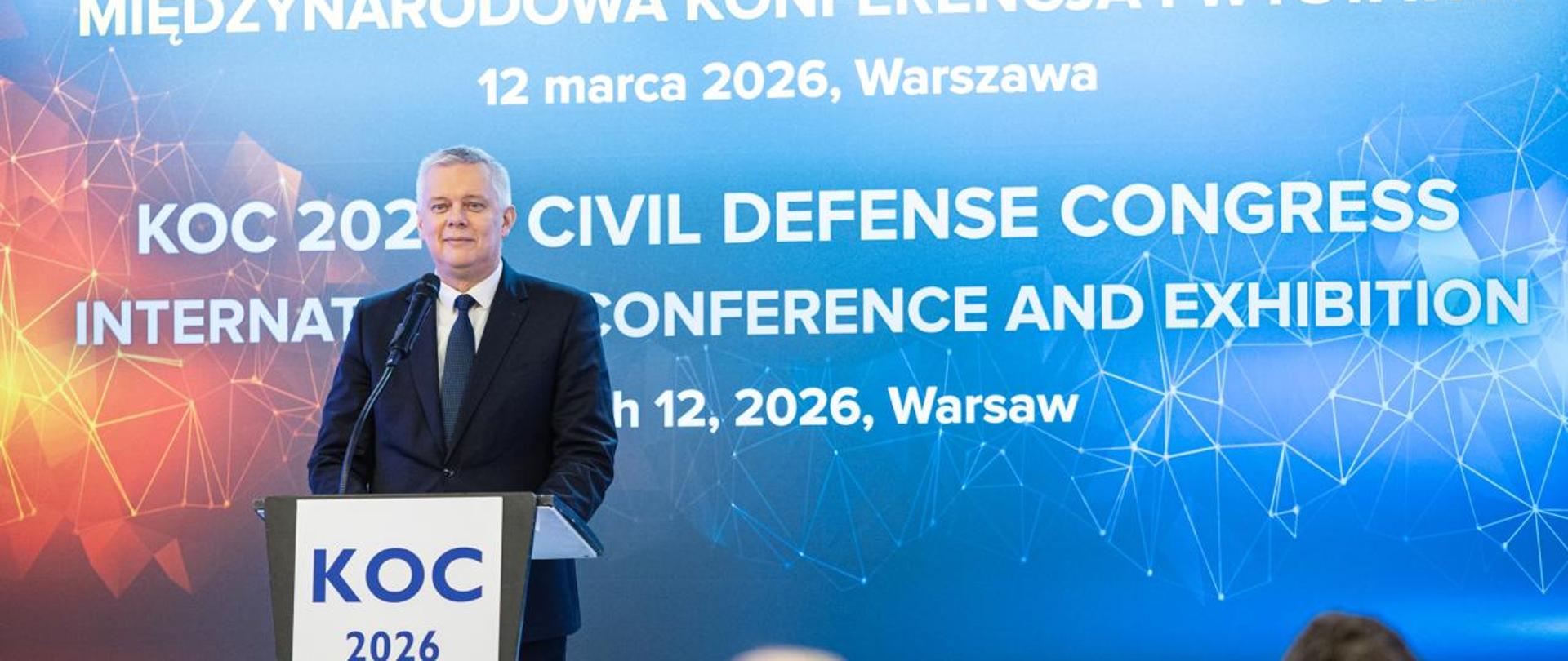 Minister Tomasz Siemoniak na scenie, przed nim biało-czarna mównica z napisem KOC 2026. W tle grafika z napisem: KOC 2026- Kongres Obrony Cywilnej Międzynarodowa Konferencja i wystawa 12 marca 2026, Warszawa.