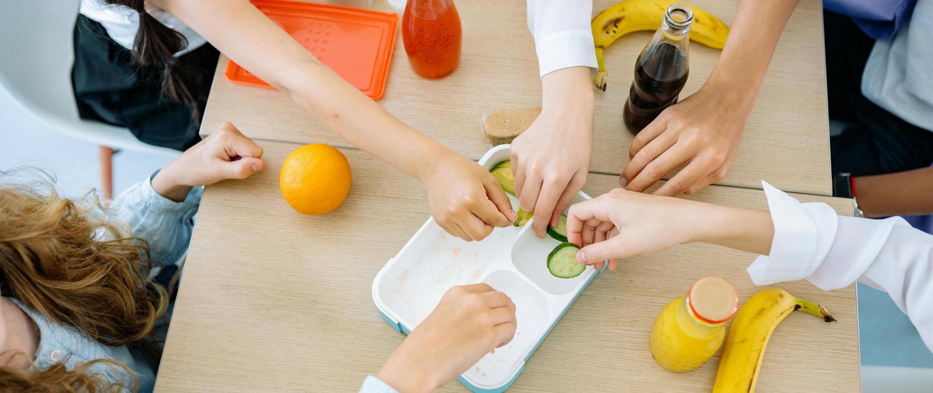 Przy stole siedzą dzieci. Nie widać ich twarzy. Widać pięć dłoni sięgających po jedzenie. Na stole znajduje się pojemnik z jedzeniem oraz na stole leżą banany, pomarańcze, jabłka i soki w szklanych butelkach