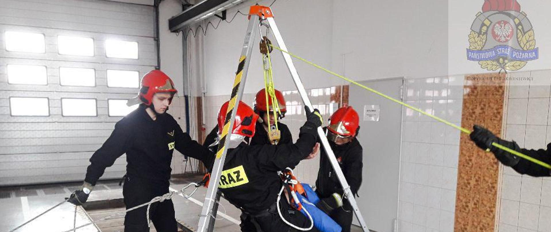 Zdjęcie przedstawia strażaków z jednostki ratowniczo-gaśniczej w skierniewicach podczas ćwiczeń z zakresu ratownictwa wysokościowego. Rozłożony jest trójnóg ratowniczy do którego za pomocą układu linowego podpięty jest strażak z osobą poszkodowaną. Ćwiczenia symulują wyciąganie osoby poszkodowanej wraz z ratownikiem ze studni, którą symuluje kanał rewizyjny do pojazdów w garażu budynku komendy. 