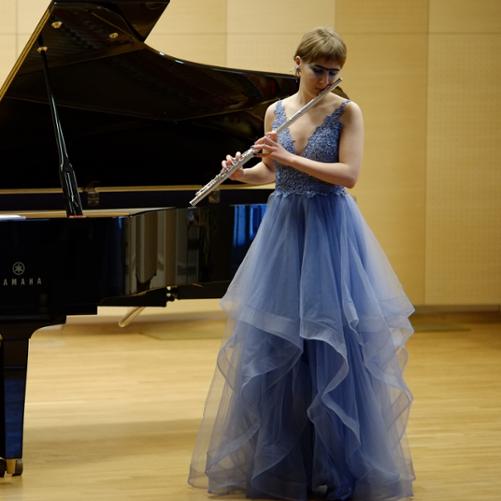 zdjęcie przedstawia uczennicę trzymającą przy twarzy flet poprzeczny oraz akompaniatora rozpoczynającego utwór na fortepianie na scenie sali koncertowej