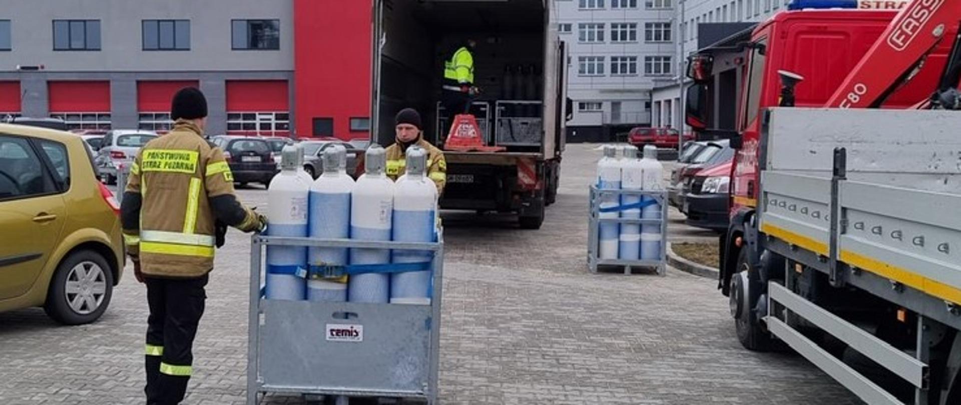 Na zdjęciu widzimy dwóch strażaków podczas rozładunku nowej dostawy butli z tlenem medycznym do banku tlenu. W tle skrzynia samochodu dostarczającego ładunek.