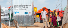 Zadowoleni Afrykańczycy w kolorowych strojach przed punktem poboru wody i latryną dla uchodźców 