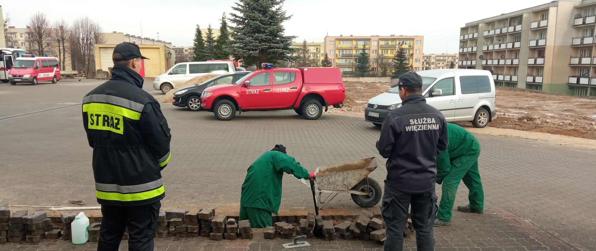 Więźniowie ubrani w zielone stroje rozbierają polbruk na placu komendy. Koło nich znajduje się taczka. Przed nimi stoją funkcjonariusz PSP ora SW. Na ziemi rozłożona jest folia ochronna.