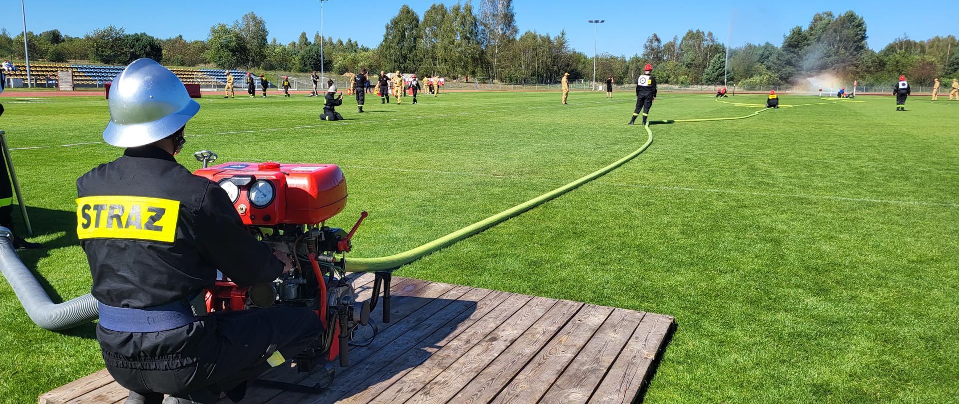 Na zdjęciu widać ćwiczenia strażackie odbywające się na zielonym boisku. Na pierwszym planie strażak w czarnym mundurze z napisem „STRAŻ” na plecach i w hełmie obsługuje przenośną motopompę ustawioną na drewnianym podeście. Do urządzenia podłączony jest wąż strażacki, który biegnie w głąb boiska. W tle kilku strażaków w czarnych mundurach i czerwonych hełmach, z końca węża tryska woda, tworząc lekką mgiełkę i tęczę. Na dalszym planie widać także osoby w jasnożółtych ubraniach oraz drzewa otaczające teren. Całość odbywa się w słoneczny dzień.