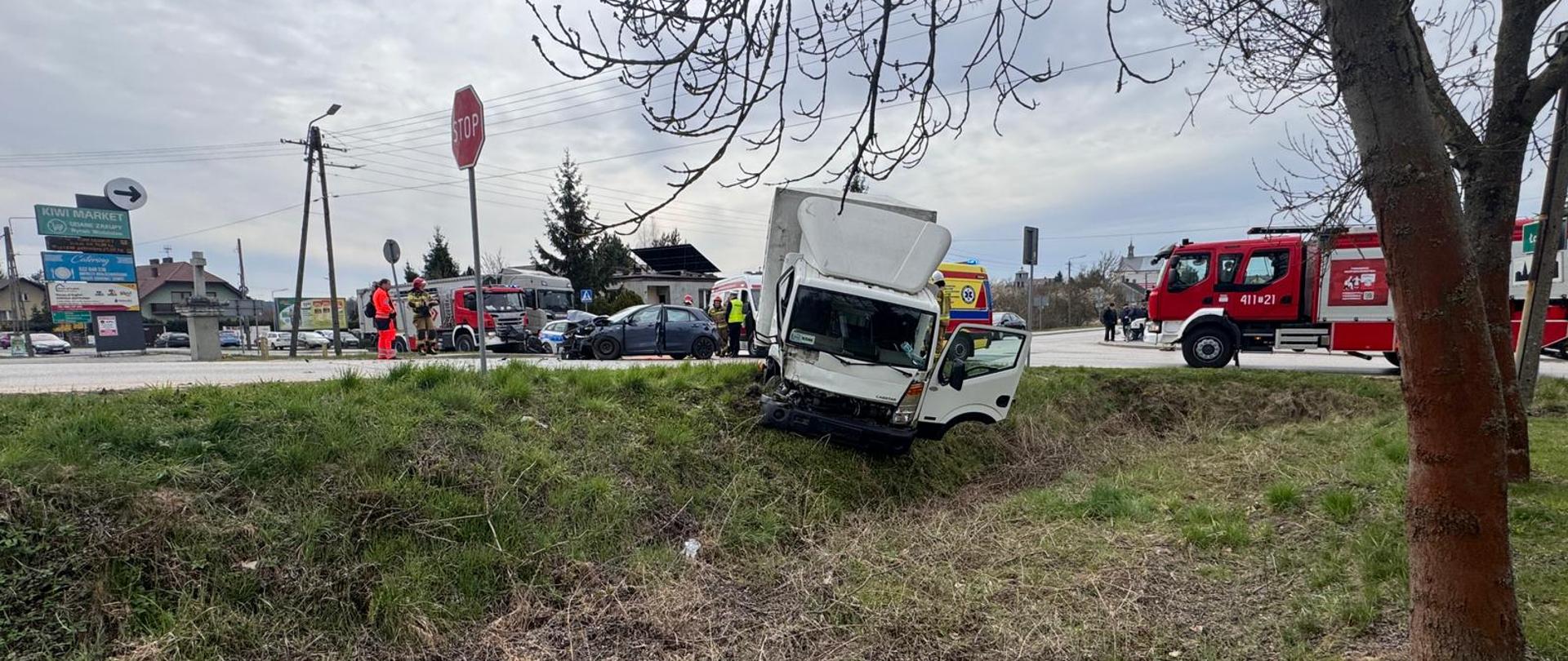 Na zdjęciu widać uszkodzony pojazd dostawczy częściowo w rowie. W tle karetka pogotowia, radiowóz policyjny, samochód ratowniczo-gaśniczy oraz pracujący funkcjonariusze straży pożarnej oraz policji.