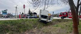 Na zdjęciu widać uszkodzony pojazd dostawczy częściowo w rowie. W tle karetka pogotowia, radiowóz policyjny, samochód ratowniczo-gaśniczy oraz pracujący funkcjonariusze straży pożarnej oraz policji.