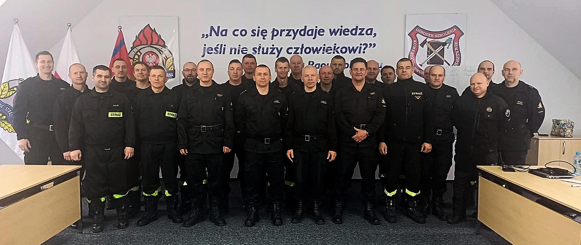 Grupa 28 strażaków - uczestników kursu nurkowego pozująca do zdjęcia grupowego. Za grupą strażaków na ścianie widoczny jest motto o treści: "Na co się przydaje wiedza jeśli nie służy człowiekowi" 
